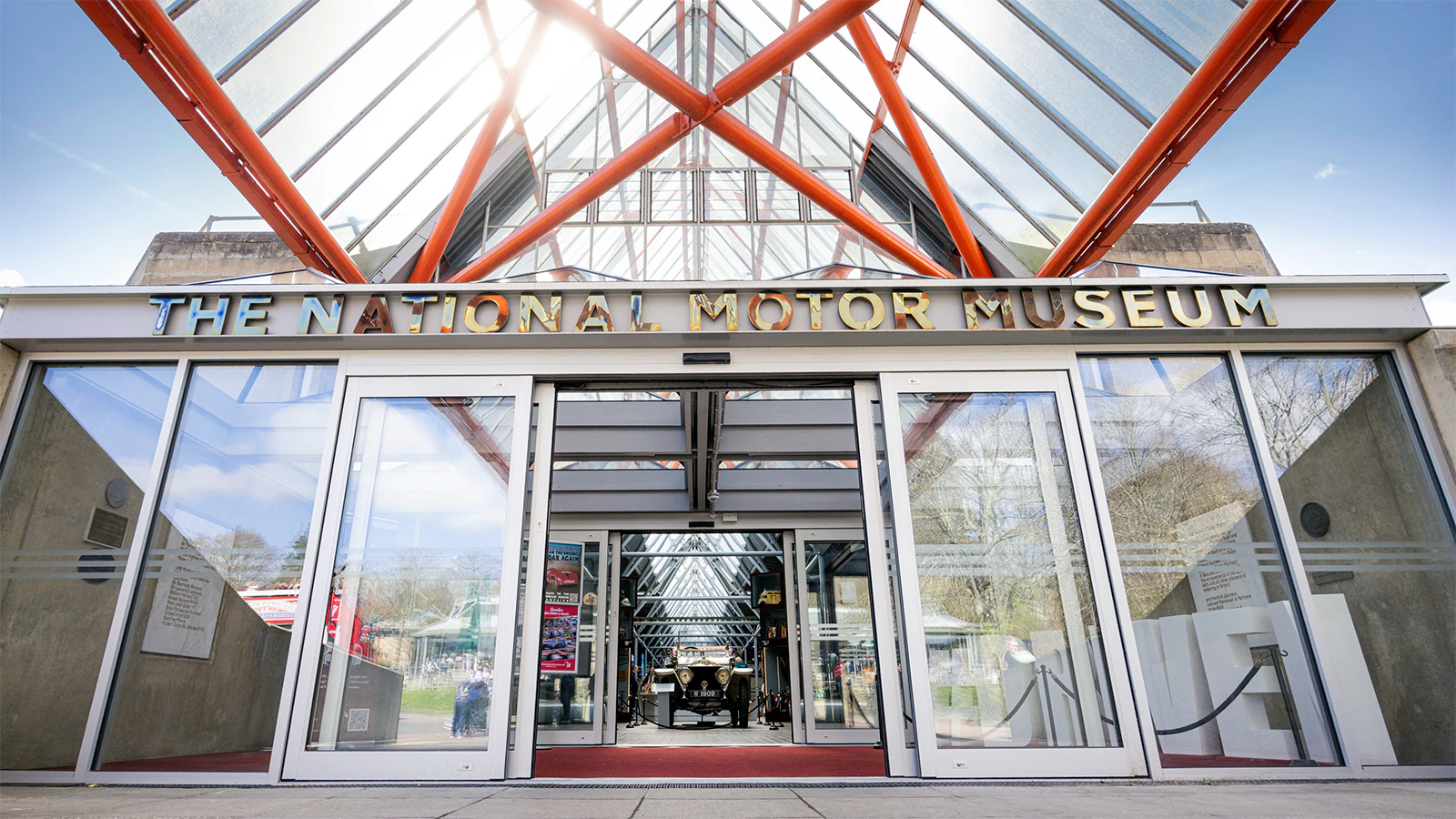 The entrance of The National Motor Museum, featuring glass doors and a peaked glass roof with orange beams overhead, seen on a bright day.
