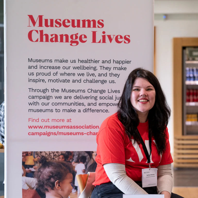 A woman in a red Museums Change Lives shirt sits and smiles beside a banner promoting the positive impact of museums on communities. The banner includes text and a link for more information.