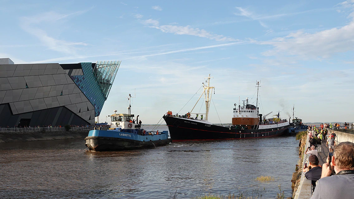 A crowd watches as two ships, including a tugboat, move along a river near a modern angular building under a blue sky with light clouds.