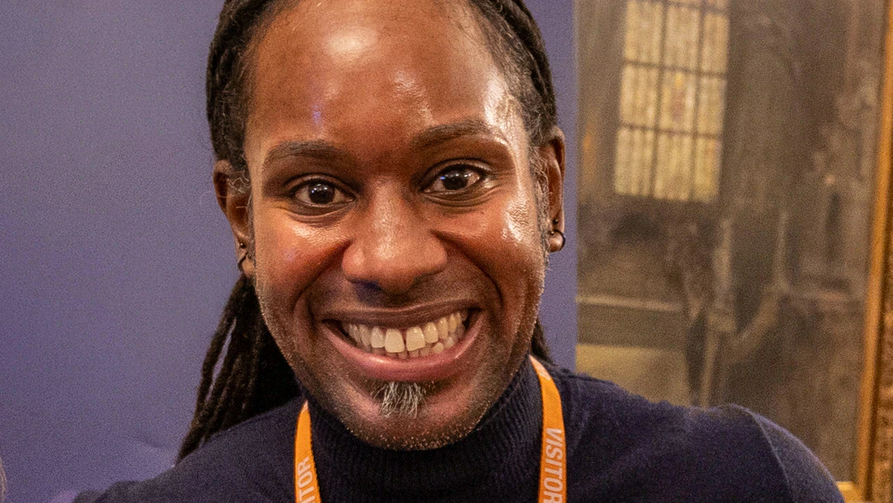 A person with long hair pulled back is smiling broadly. They are wearing a dark sweater and an orange lanyard.