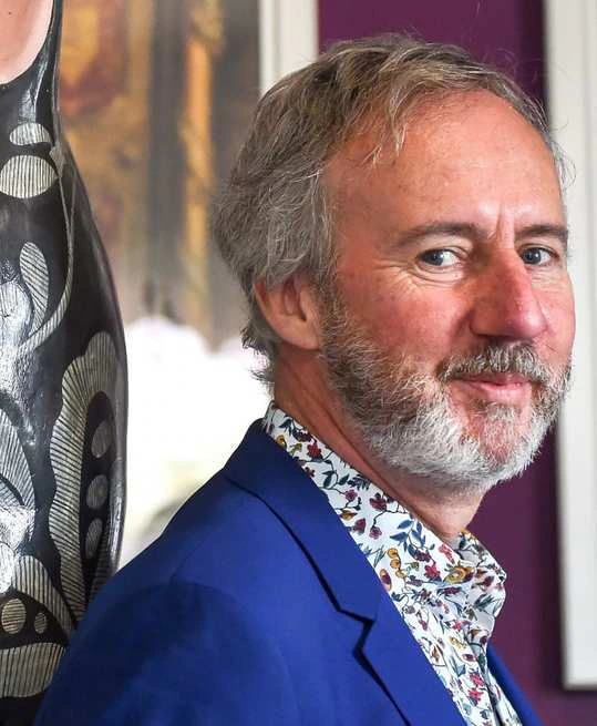 A man with gray hair and a beard, wearing a blue blazer and a patterned shirt, stands indoors and looks toward the camera, smiling slightly. Theres patterned artwork and a blurred background behind him.