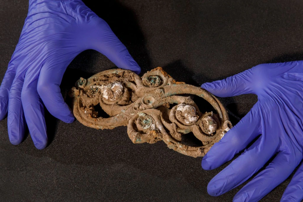 Two gloved hands carefully hold an ornate, corroded metal artifact with intricate swirl patterns and some white encrustations, against a dark surface.