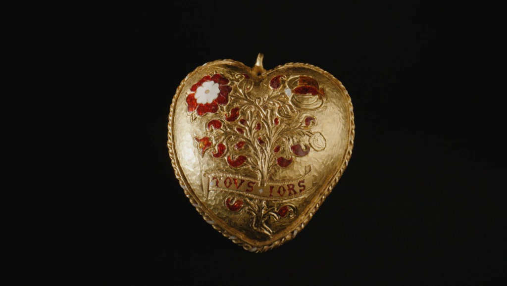 A gold, heart-shaped locket with a raised tree design, red and white flowers, and the word LOVS: TORS engraved, set against a black background.