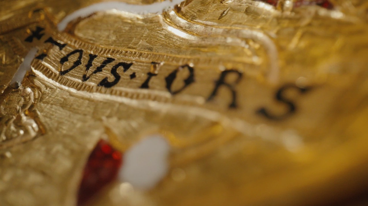 Close-up of a golden object with raised letters spelling TOUSIORS in black, surrounded by engraved patterns and red and white decorations. The image has a shiny, reflective texture.