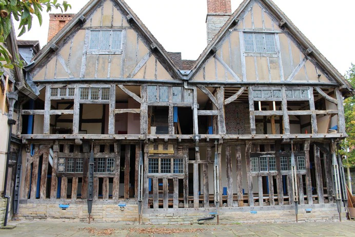 A historic timber-framed house with missing walls and exposed beams, revealing the interior rooms and structure. The building appears to be undergoing restoration or repair.