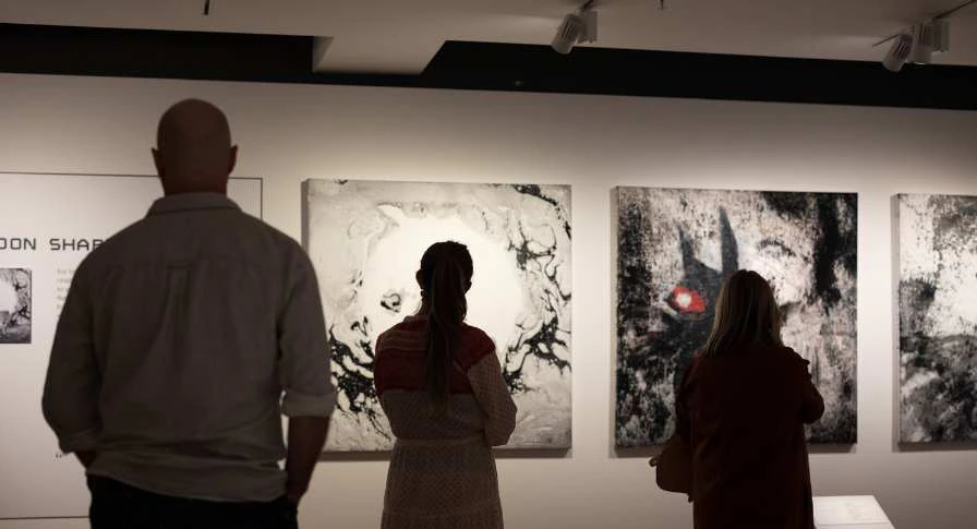 Three people stand in an art gallery viewing abstract black and white paintings, one with a red accent, displayed on a well-lit wall.
