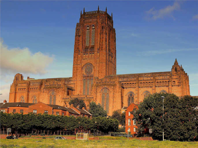 A large, brown stone cathedral with a tall central tower rises above trees and red-brick houses under a blue sky with scattered clouds.