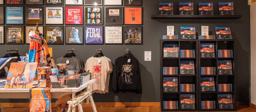 A gift shop with shelves of neatly stacked books, T-shirts, hoodies, scarves, and art prints on the wall above. The floor is wooden and the walls are gray.