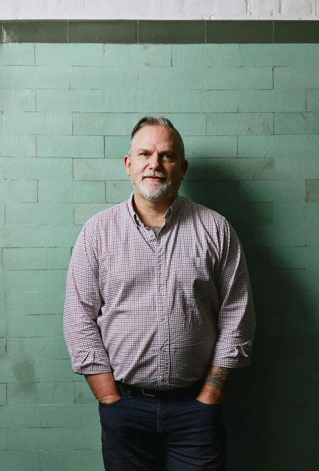 A middle-aged man with gray hair and a beard stands against a green tiled wall, wearing a checked shirt with sleeves rolled up and dark pants, looking at the camera with a neutral expression.
