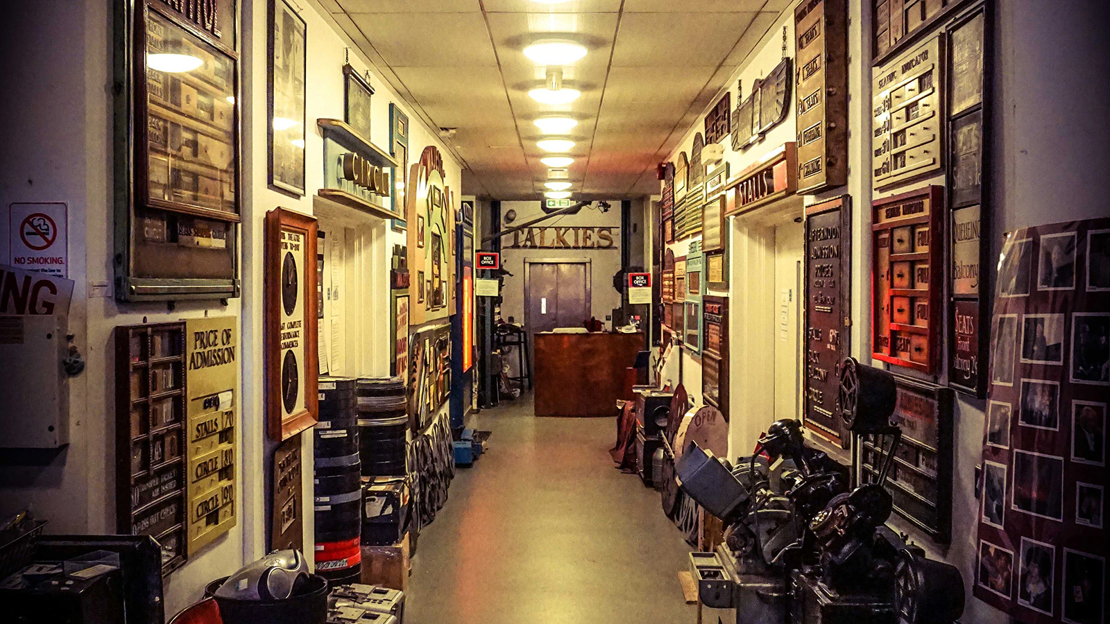 A hallway lined with vintage film equipment, posters, and memorabilia, leading to a wood desk at the end under bright ceiling lights. Walls are densely decorated with photos and signs.