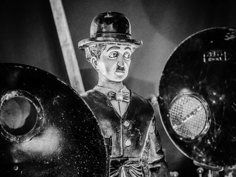 Black and white photo of a Charlie Chaplin figurine wearing a bowler hat and bow tie, standing between two vintage film lights.
