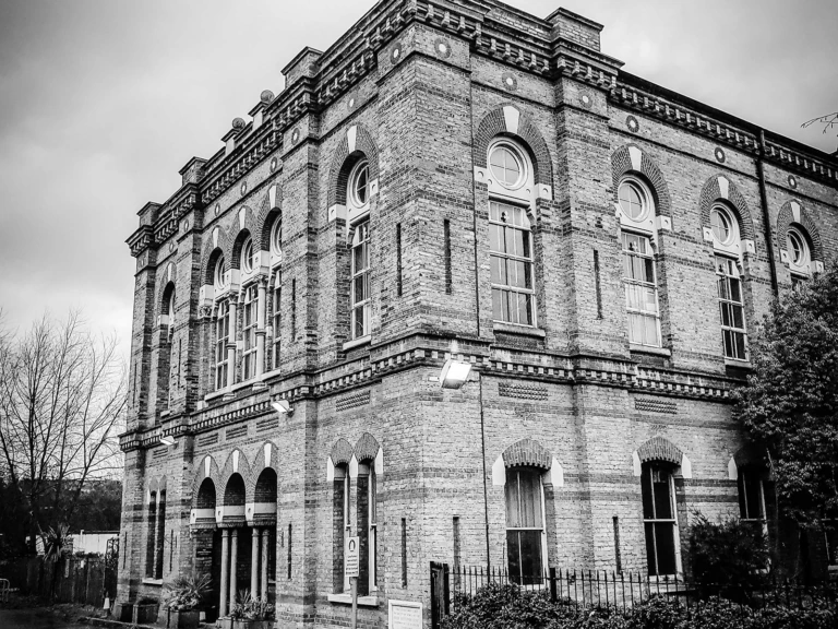 A black and white photo of a large, old brick building with arched windows, decorative details, and a fenced garden. Leafless trees are on the left, and the sky is cloudy.