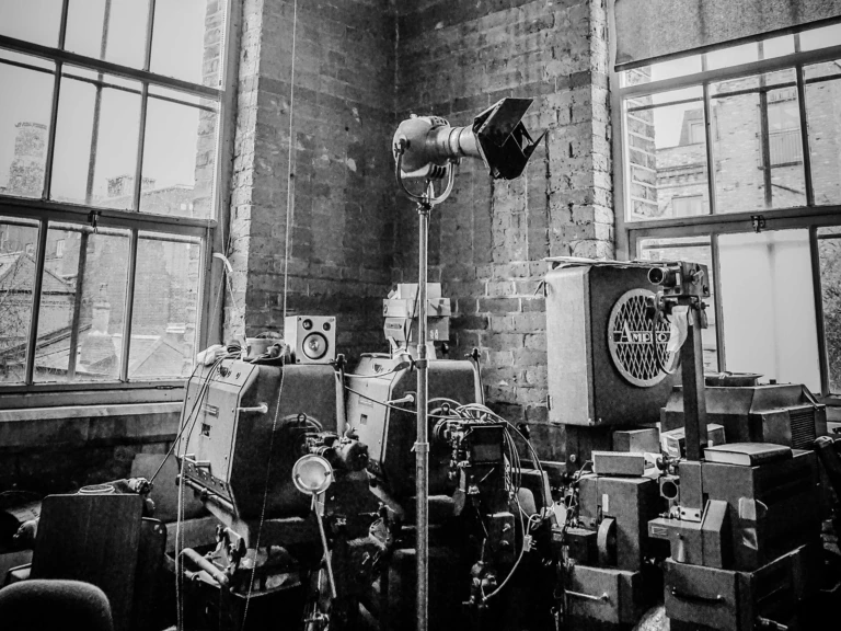 Black and white photo of a cluttered industrial room with large machinery, wires, control panels, and a light fixture. Two large windows reveal brick buildings outside.