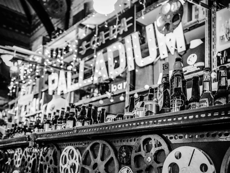 Rows of beer bottles line a bar, with movie reels and string lights as decor. A large PALLADIUM sign hangs in the background. The setting is lively and eclectic in black and white.