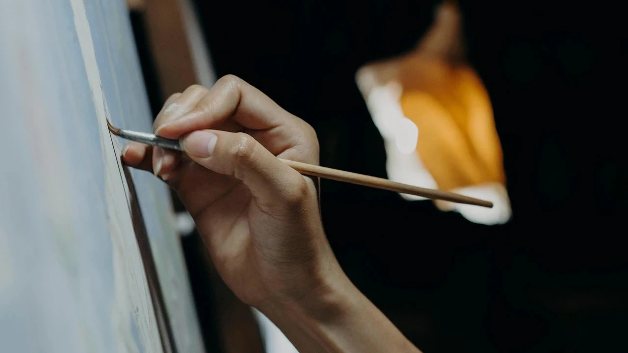 A persons hand holding a paintbrush, painting on a canvas with soft lighting and blurred background.