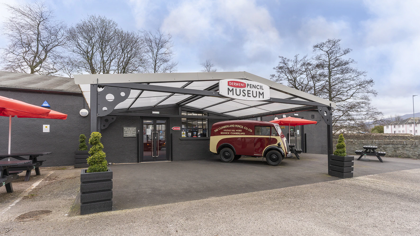 The entrance of the Derwent Pencil Museum features a vintage red and cream delivery van, black picnic tables with red umbrellas, and potted plants by the doors on a cloudy day.