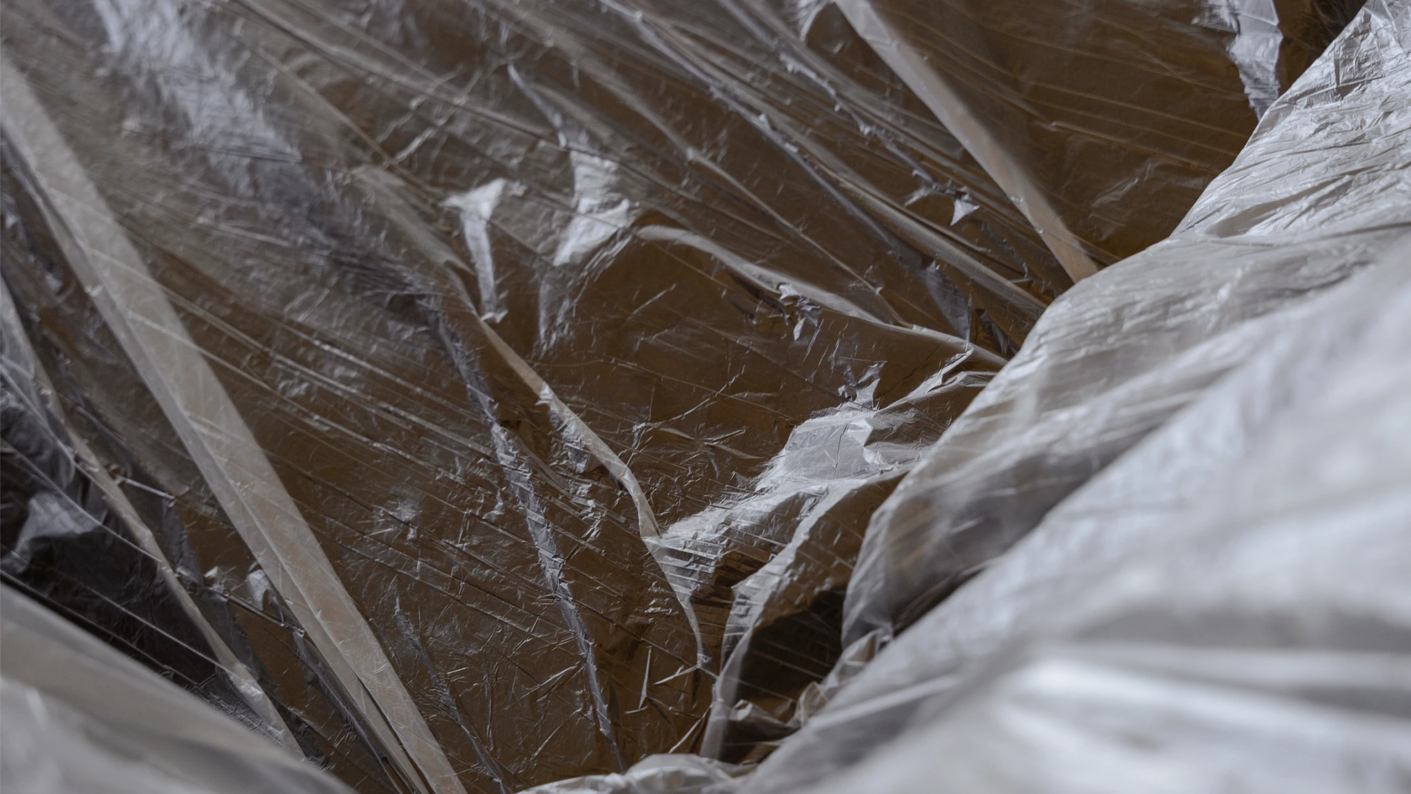 Close-up of wrinkled, transparent plastic sheeting covering an unknown brown surface, with reflections and creases creating abstract patterns.