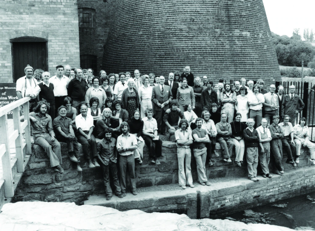 A large group of people pose together outside in front of a brick building with a large, conical kiln structure. Some stand on steps, while others sit or lean on a stone wall, all facing the camera.
