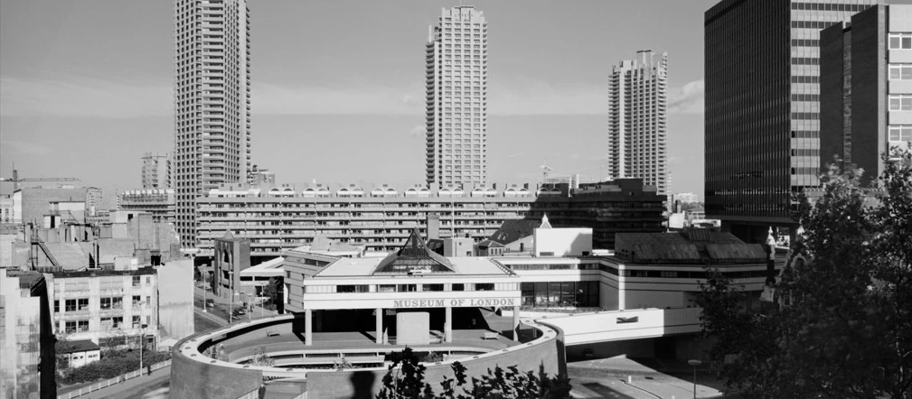 Black and white photo of Londons Barbican Estate, with three tall residential towers, the Museum of London in front, and modern office buildings surrounding the area.