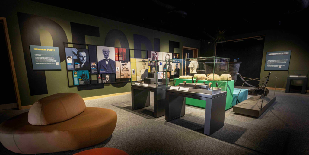 A museum exhibit room displays historical photographs, artifacts in glass cases, and interpretive signs. Sofas are arranged for seating, and the word PEOPLE is featured prominently on a wall in the background.