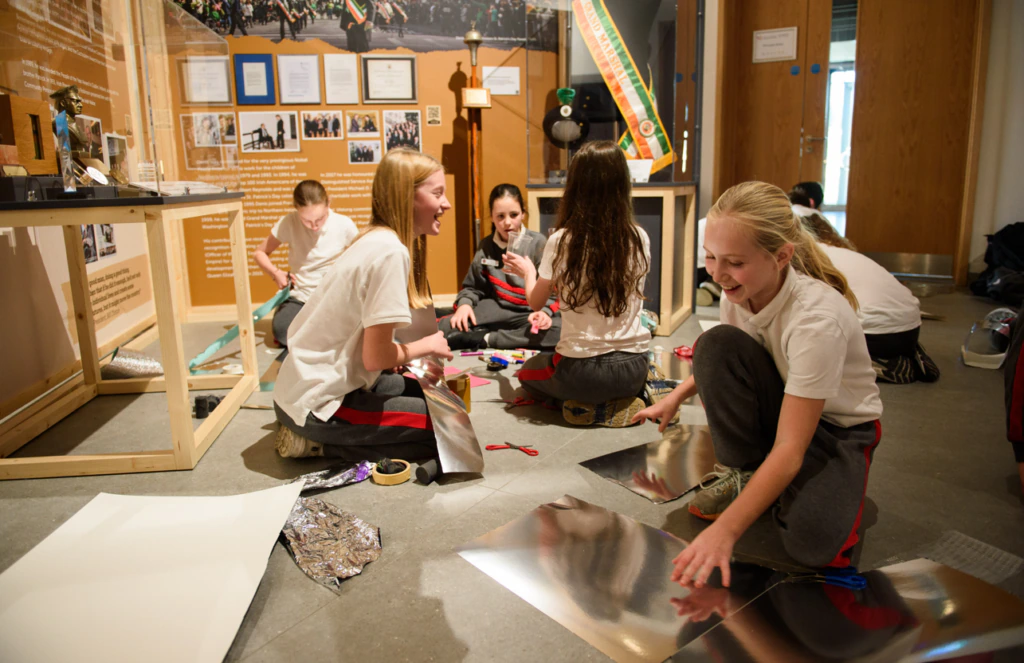 A group of girls in school uniforms sit on the floor, laughing and working on a craft project with paper, foil, and tape in a brightly lit classroom or museum setting.