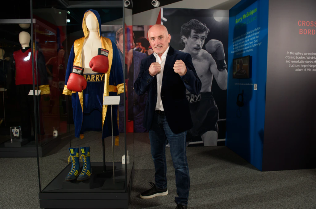 A man stands in a museum exhibit with boxing memorabilia, including gloves, boots, and a robe in a glass case. He poses with fists raised in front of a wall with boxing photos and text.