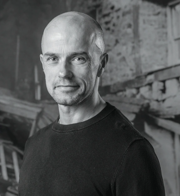 A bald man wearing a dark sweater smiles slightly while standing in front of a rustic stone building. The image is in black and white.