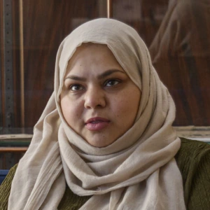A woman wearing a beige head covering and olive green dress sits on a blue couch