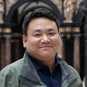 A person wearing a green jacket and dark shirt smiles while standing indoors in a museum or gallery, with classical sculptures and detailed architectural backgrounds visible behind them.