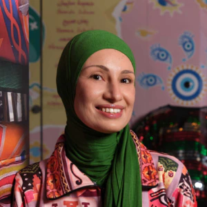 A woman wearing a bright green headscarf and a colorful patterned jacket smiles at the camera, standing in front of a colorful background