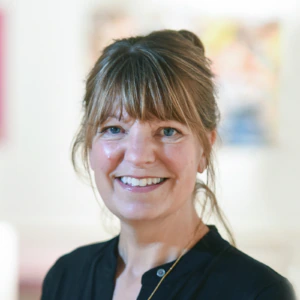 A woman with light brown hair in a loose bun and bangs smiles at the camera. She is wearing a black top and a necklace. The background is softly blurred with light and colorful shapes.