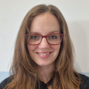 A woman with long brown hair and red glasses smiles at the camera. She is wearing a black top and is posed against a light-colored background.