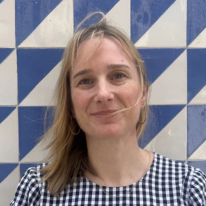 A woman with blonde hair, wearing a black and white checkered top, stands in front of a blue and white geometric tiled wall, smiling slightly.