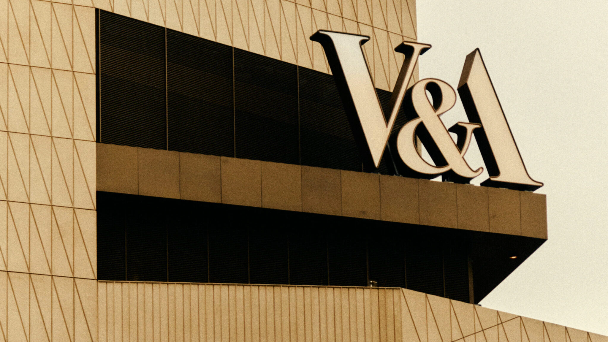 Large V&A letters are mounted on the exterior of a modern, geometric building with a patterned facade, likely representing the Victoria and Albert Museum. The image has a sepia tone, giving it a vintage feel.