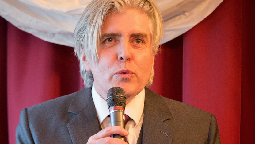 A man with gray hair in a suit speaks into a microphone at a podium, standing in front of red curtains and a white decorative fabric.