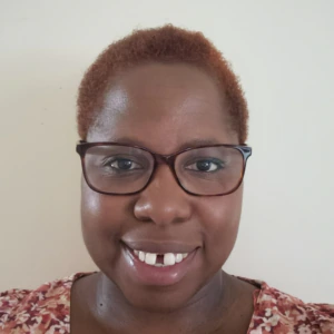 A woman with short, reddish-brown hair and glasses smiles at the camera. She is wearing a patterned top and is in front of a plain, light-colored background.