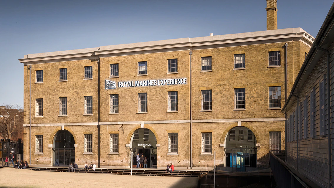 A large brick building with arched doorways and windows houses the Royal Marines Experience. People walk near the entrance, and a calm body of water is visible in the foreground.