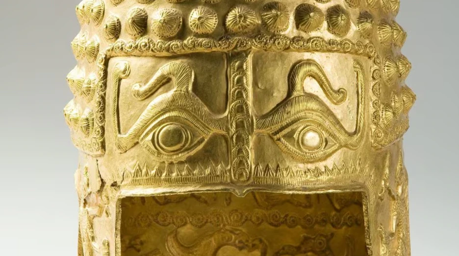 Golden ancient helmet with large engraved eyes and decorative patterns, featuring raised circular studs and intricate designs above and between the eyes.