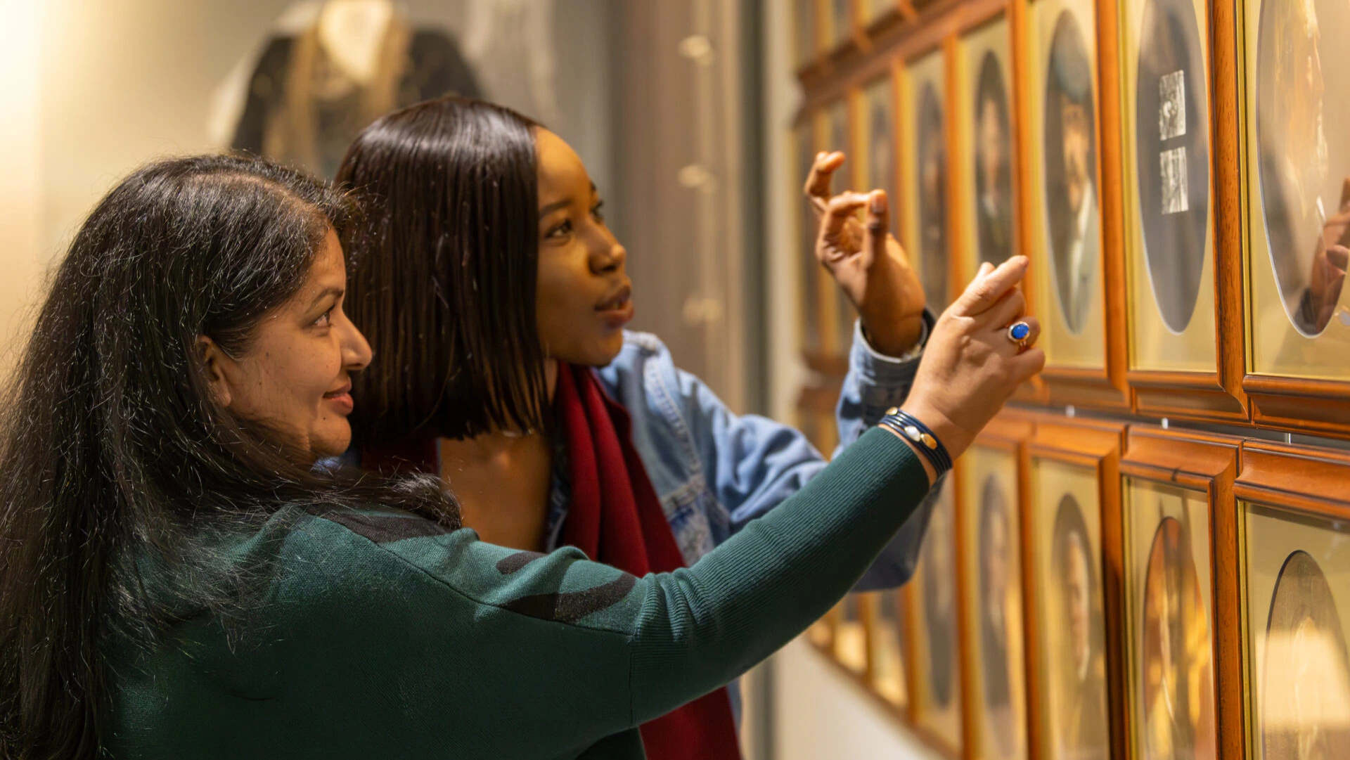 Two women look closely at framed photographs on a wall in a museum or gallery, pointing and discussing the images together.