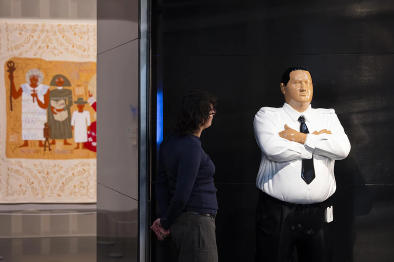A person stands next to a large, realistic statue of a man in a white shirt and black tie with arms crossed; colorful artwork hangs on a wall in the background.