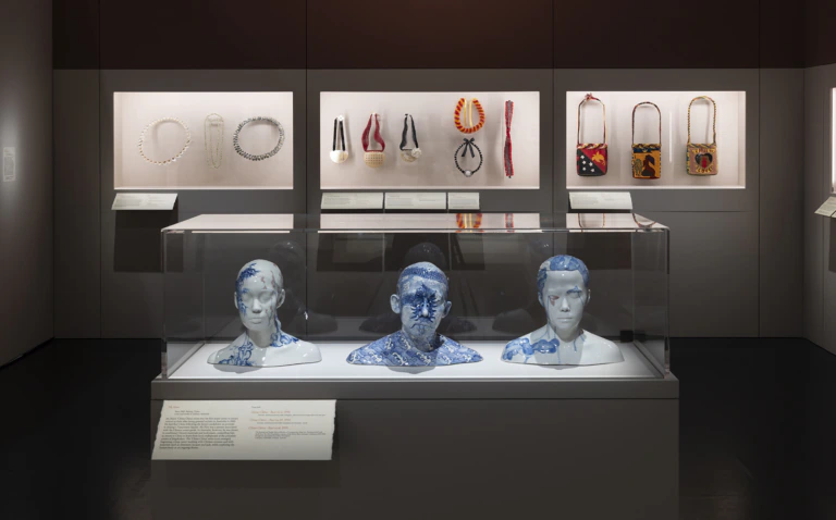 A museum display with three blue-patterned busts in a glass case and various beaded necklaces and bags on the wall behind them, each presented individually with descriptive placards.