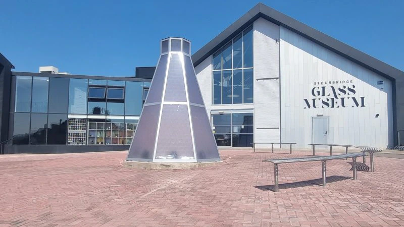 Modern building with Stourbridge Glass Museum on the wall, a glass cone sculpture in front, surrounded by benches and brick pavement under a clear blue sky.