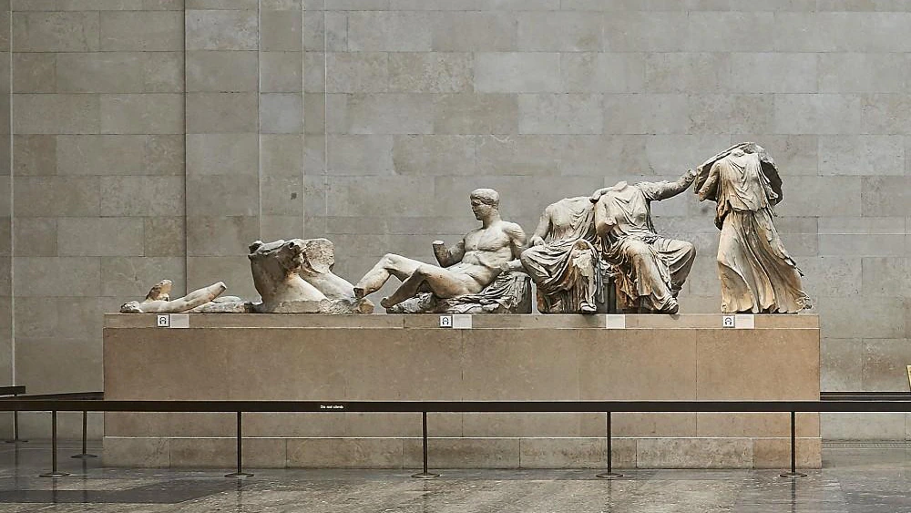 White marble statues from ancient Greece, partially damaged, are displayed on a stone platform in a museum with gray stone walls and floors. The figures are reclining or seated, missing some heads and limbs.