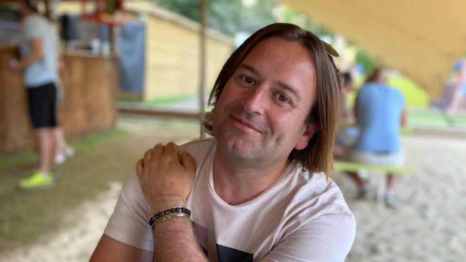 A person with medium-length brown hair sits at a blue picnic table under a canopy, smiling, with their arms crossed on the table. Other people and a wooden food stand are visible in the background.