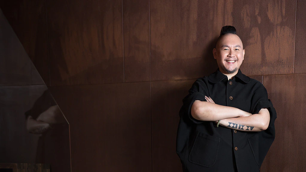 A smiling person with a short mohawk and a tattoo on their forearm stands with arms crossed, wearing a black button-up shirt, in front of a brown textured wall.