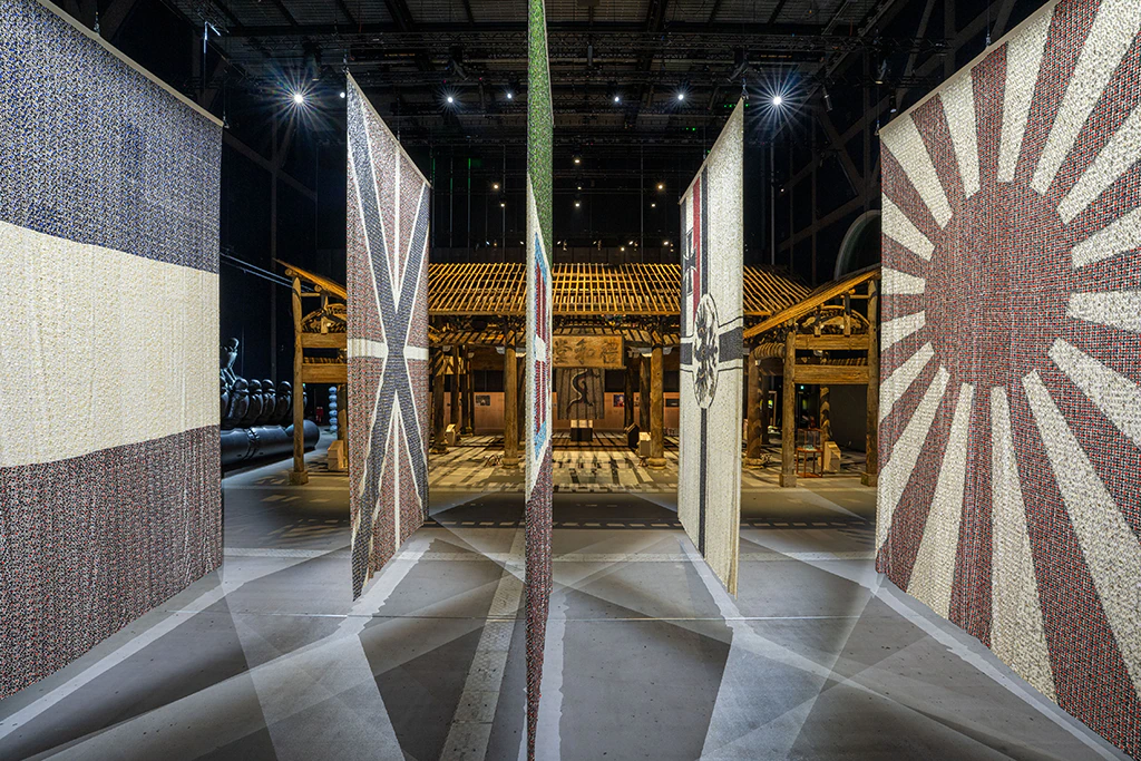Four large hanging banners with various national flag designs stand upright in an industrial space, with stage lights illuminating them and a wooden structure visible in the background.