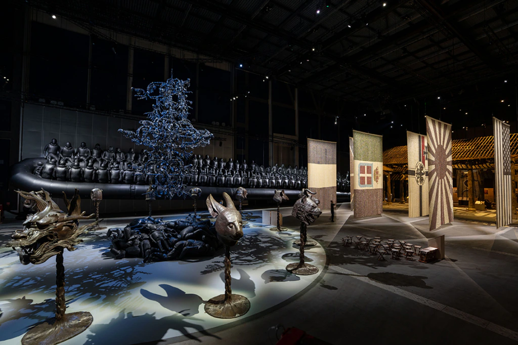 Art installation in a large hall with dragon and animal head sculptures, patterned banners, a black inflatable raft with human figures, and wooden benches under dramatic lighting.