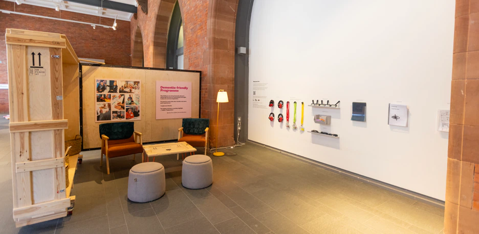 A museum exhibit area with two chairs, a lamp, ottomans, an informational display, and various objects mounted on the wall, including headphones and colorful tactile items. Brick arches and shipping crates are visible.