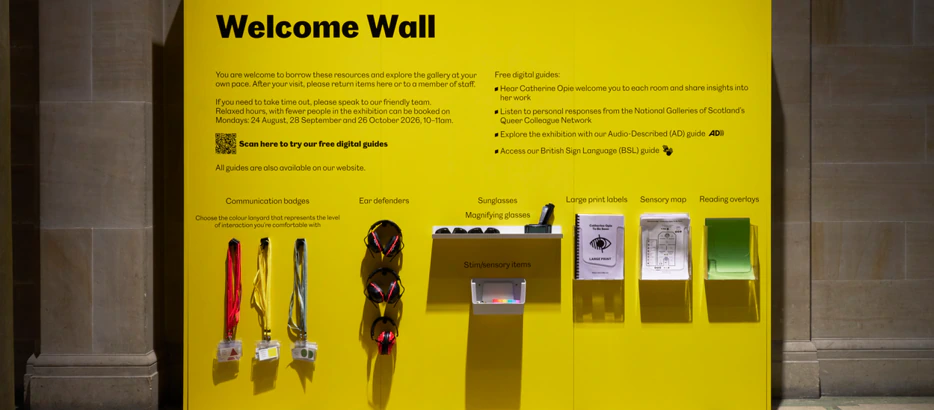 A bright yellow Welcome Wall displays information, guides, communication badges, sensory items, large print labels, a sensory map, and a British Sign Language leaflet in a museum or gallery setting.