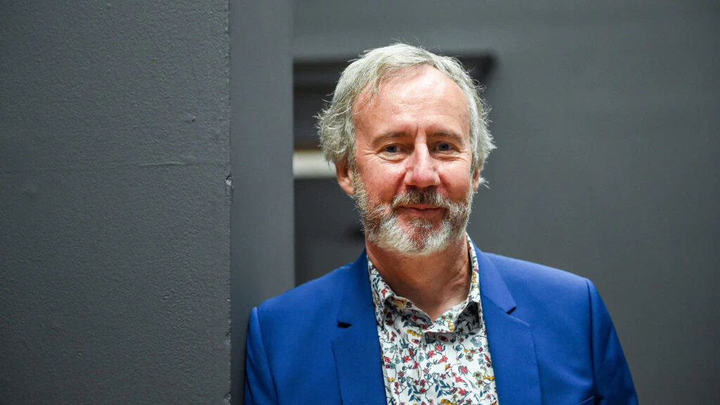 A man with gray hair and a beard, wearing a blue blazer and a floral shirt, stands in front of a dark gray wall, smiling slightly at the camera.